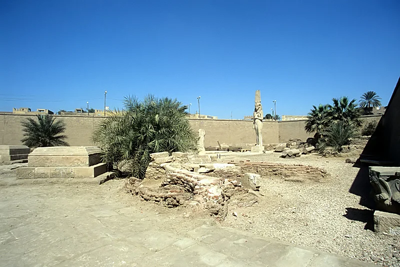 An open-air enclosure of ancient stone blocks and a standing statue among palms under a blue sky.
