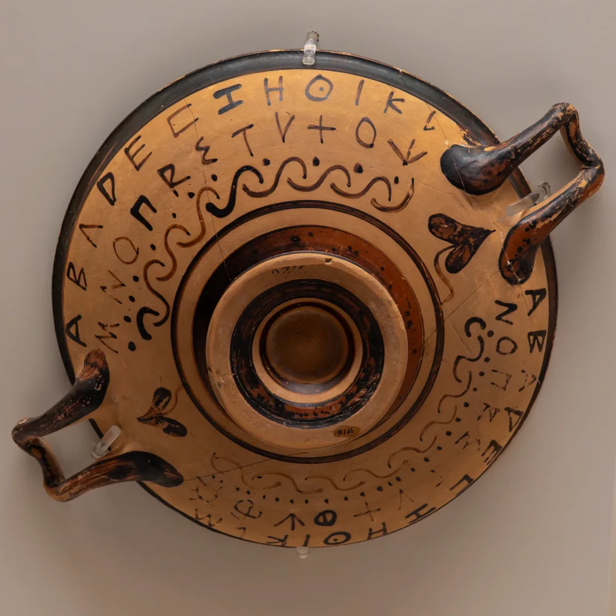 A shallow two-handled cup seen from above, the Greek alphabet painted in black letters round its rim.