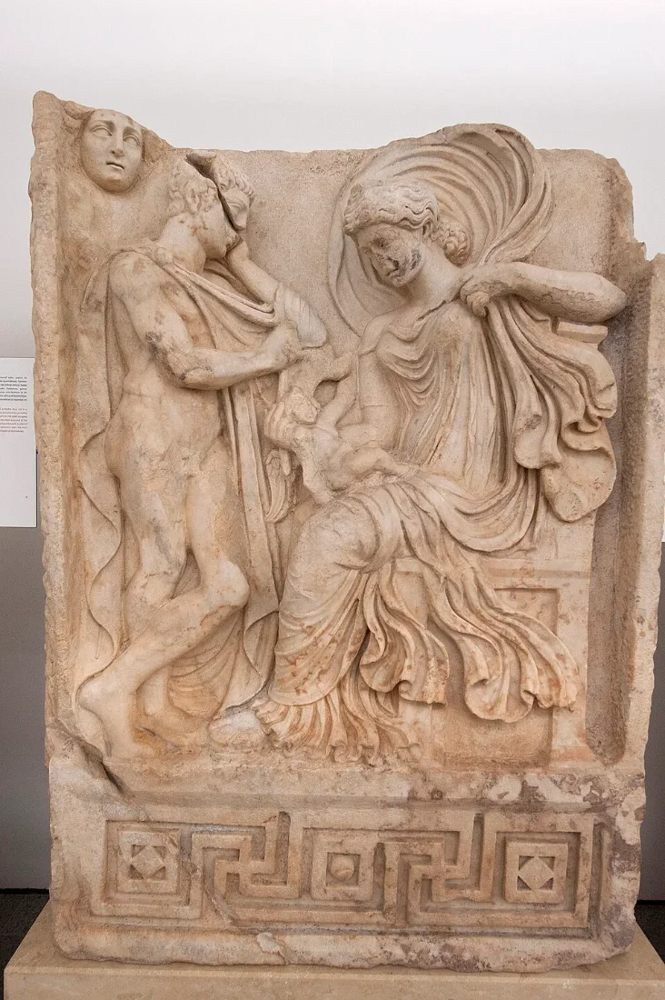 A marble relief of a seated man and a woman whose cloak billows over them, with a small winged figure.