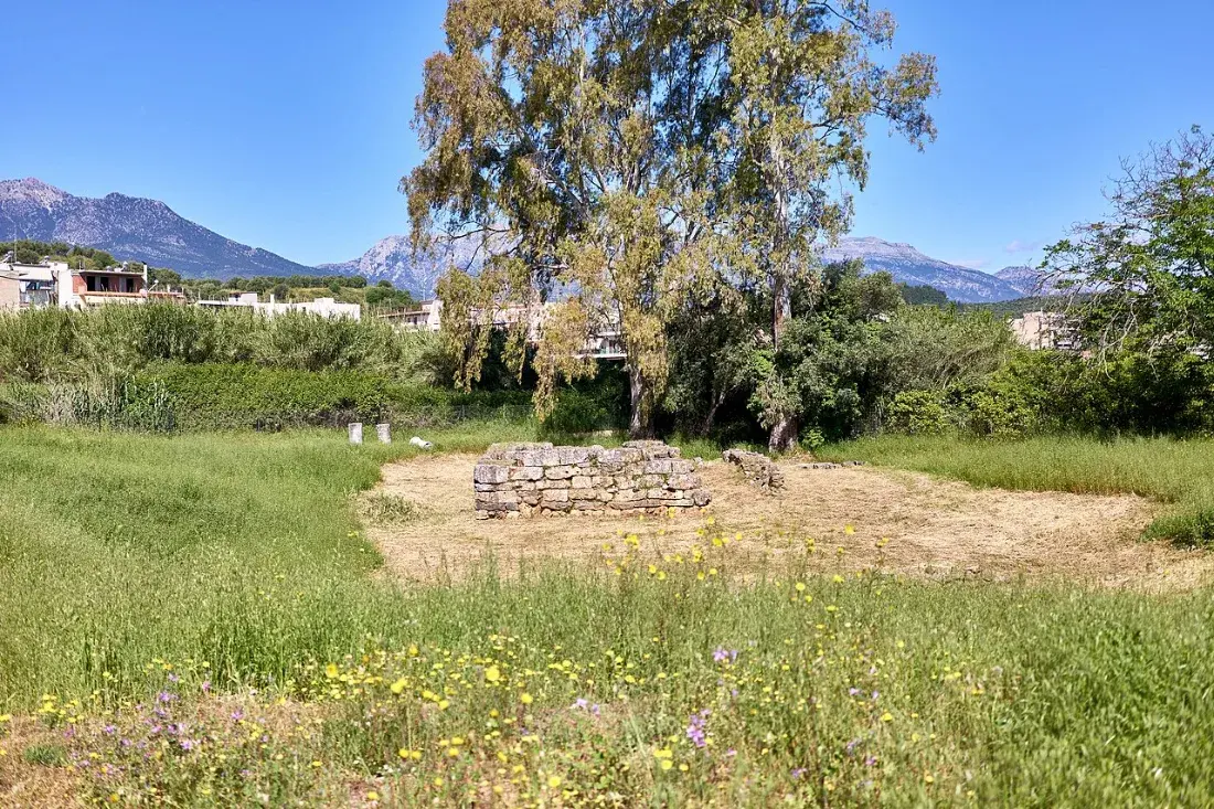 Low ruined walls in a grassy field, mountains behind.