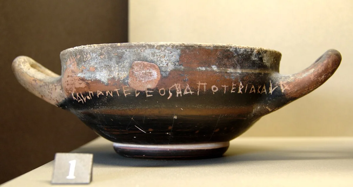 A black-glazed two-handled drinking cup with a line of incised Greek letters around its body.