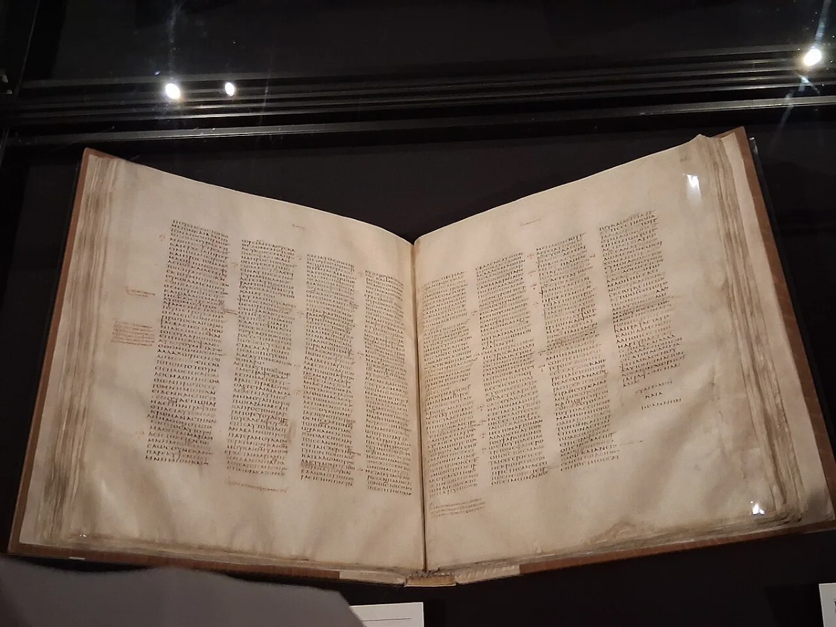 An open parchment codex lying in a glass display case, its two pages written in four columns each.