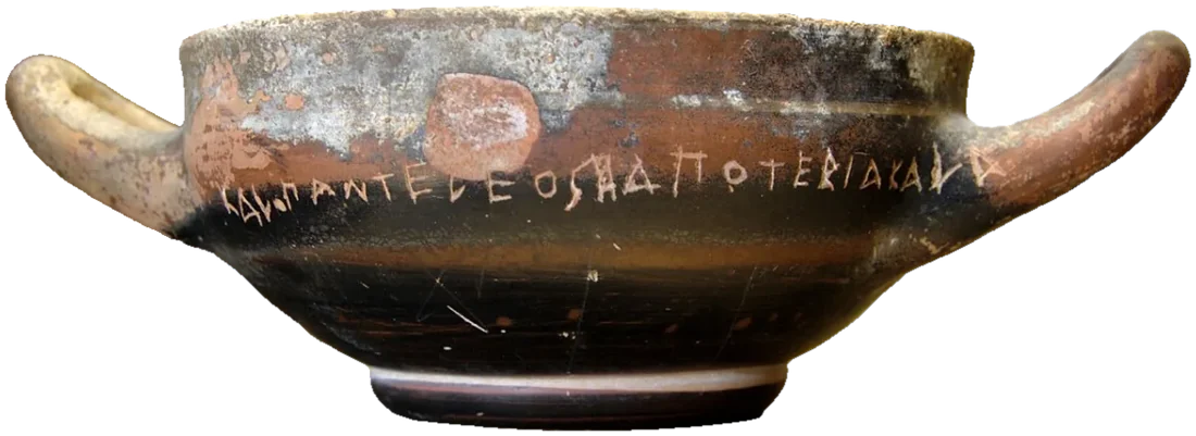 A black-glazed two-handled drinking cup with a line of incised Greek letters around its body.