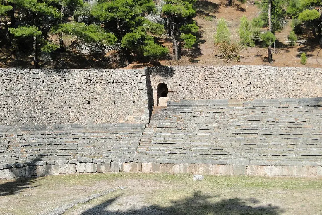 Stone seating rising along a long racetrack under pine trees.