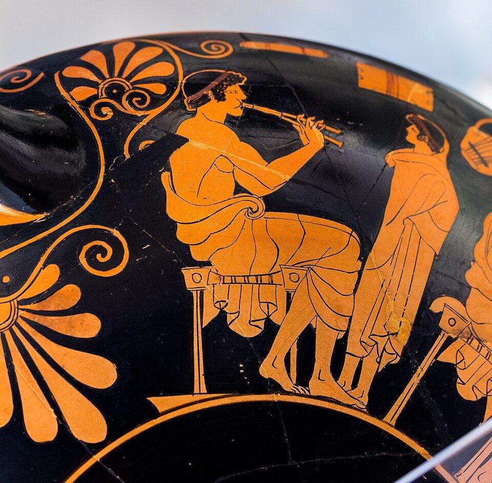 Red-figure detail: a seated man plays the double pipes while a boy stands before him.