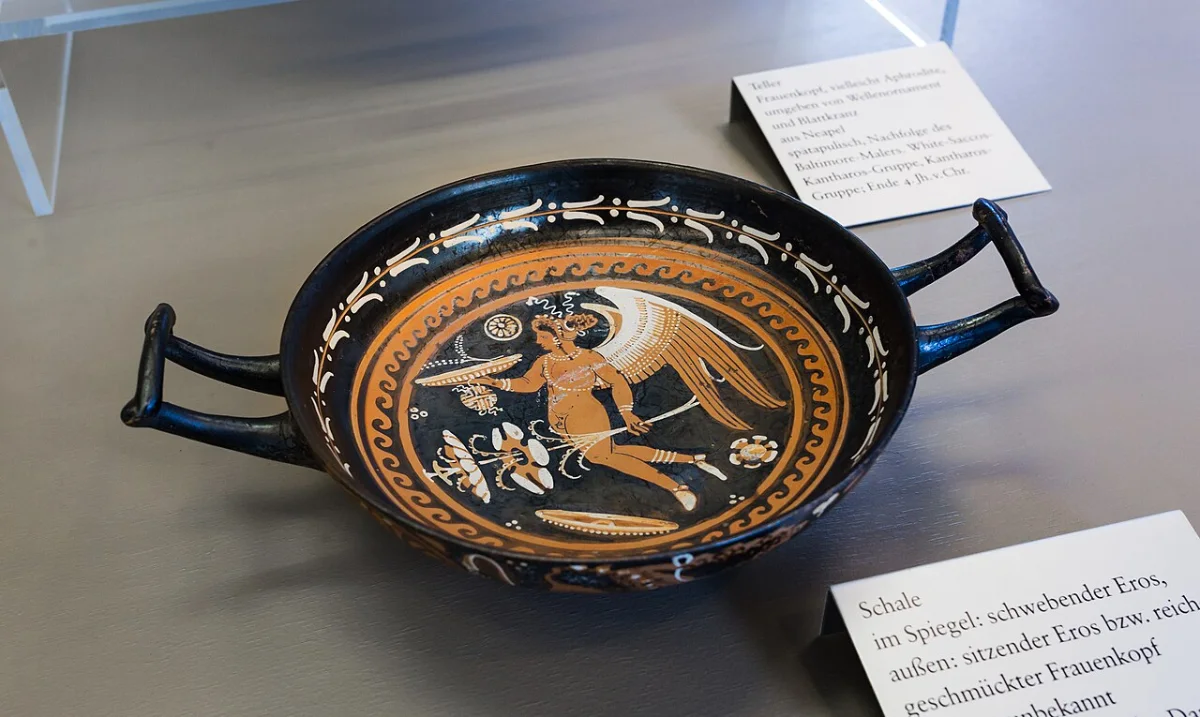 A red-figure cup seen from above: inside, a winged Eros flies holding a dish, a ball and a plant.