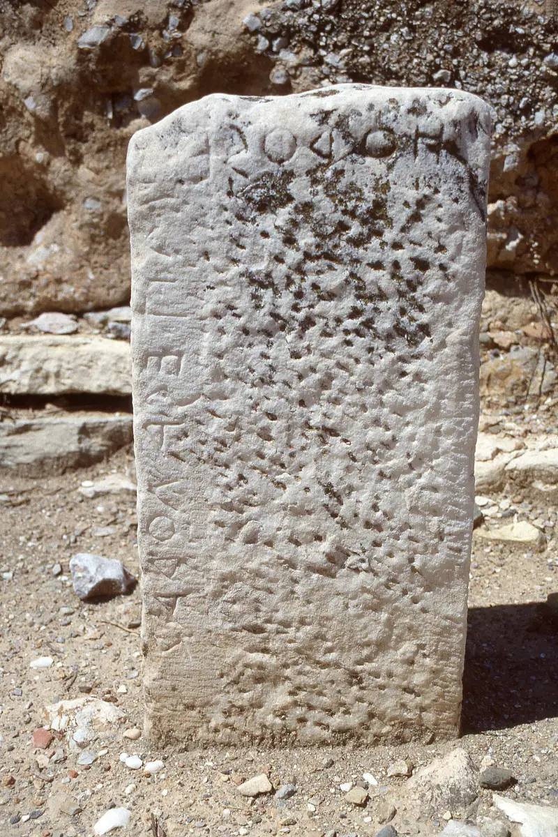 A worn upright marble post inscribed near its top with Greek letters, standing in the ground.
