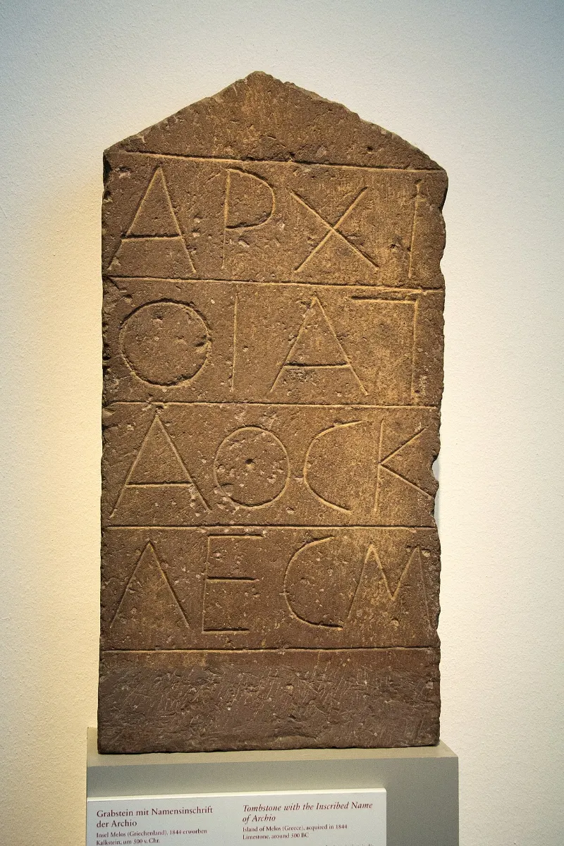 A narrow limestone gravestone with a pointed top, carved with large archaic Greek letters in short lines.
