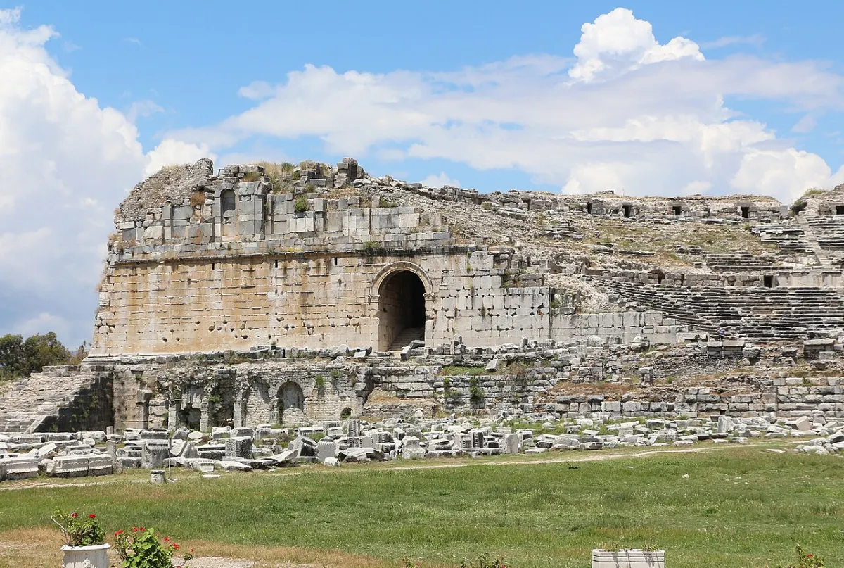 The great curved stone wall and arched entrance of an ancient theatre in a grassy field.
