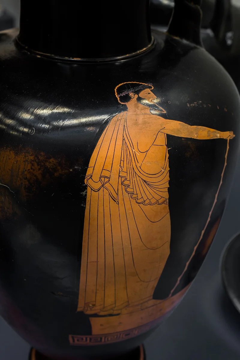 Red-figure amphora: a bearded man in a cloak stands on a low platform, one arm stretched out, words issuing from his mouth.
