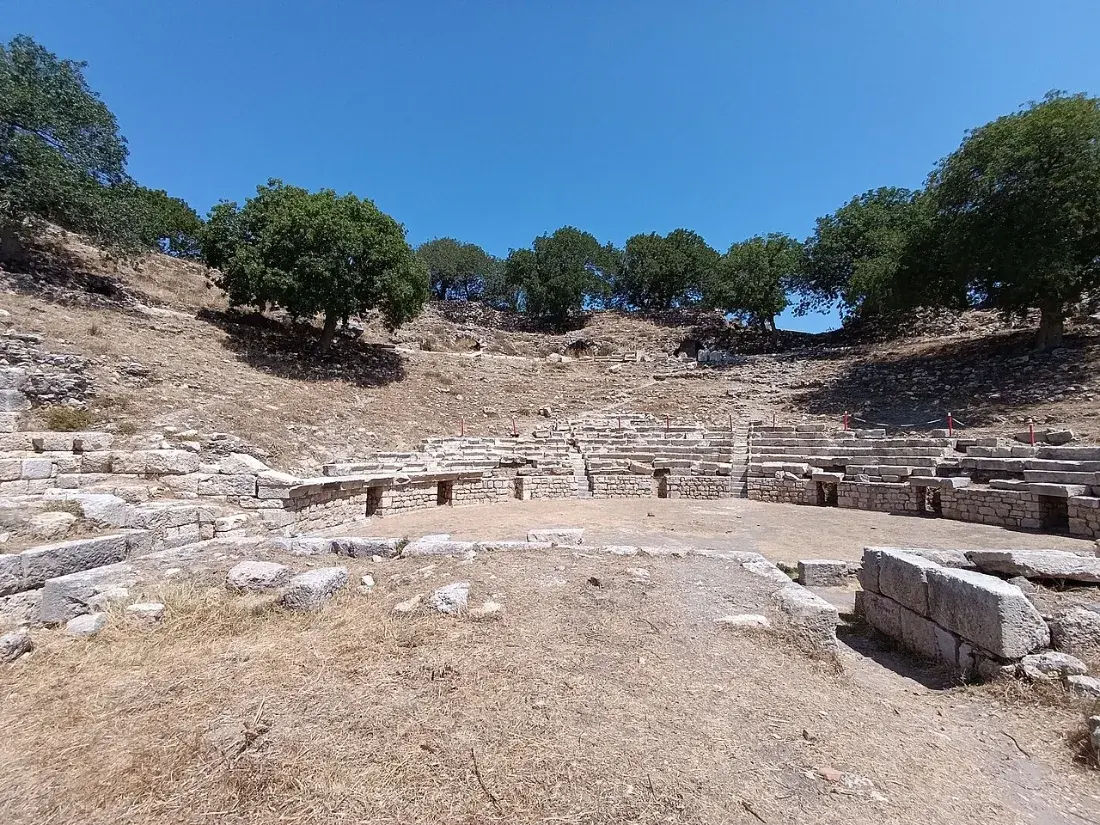 The stone seats of a small theatre among olive trees.