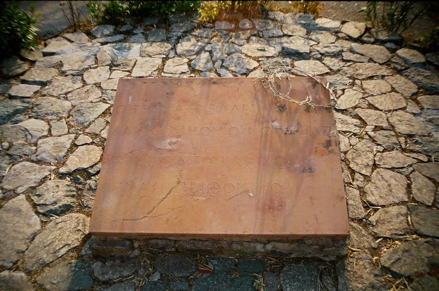 A stone slab carved with lines of Greek, set in a paved area among plants.