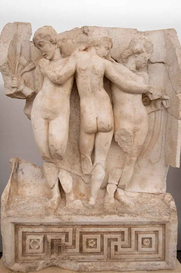 A marble relief of three naked women embracing, the middle one seen from behind.